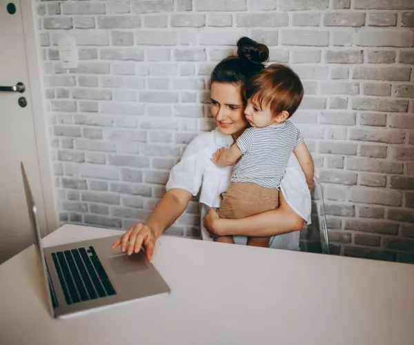 Woman at work on laptop holds an infant as she types